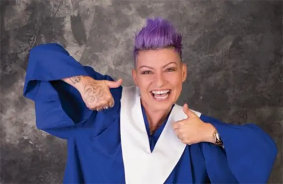 Sabrina smiles in grad gown, happy to be accepted as a Hospital Support Specialist, purple hair and tattoos included!